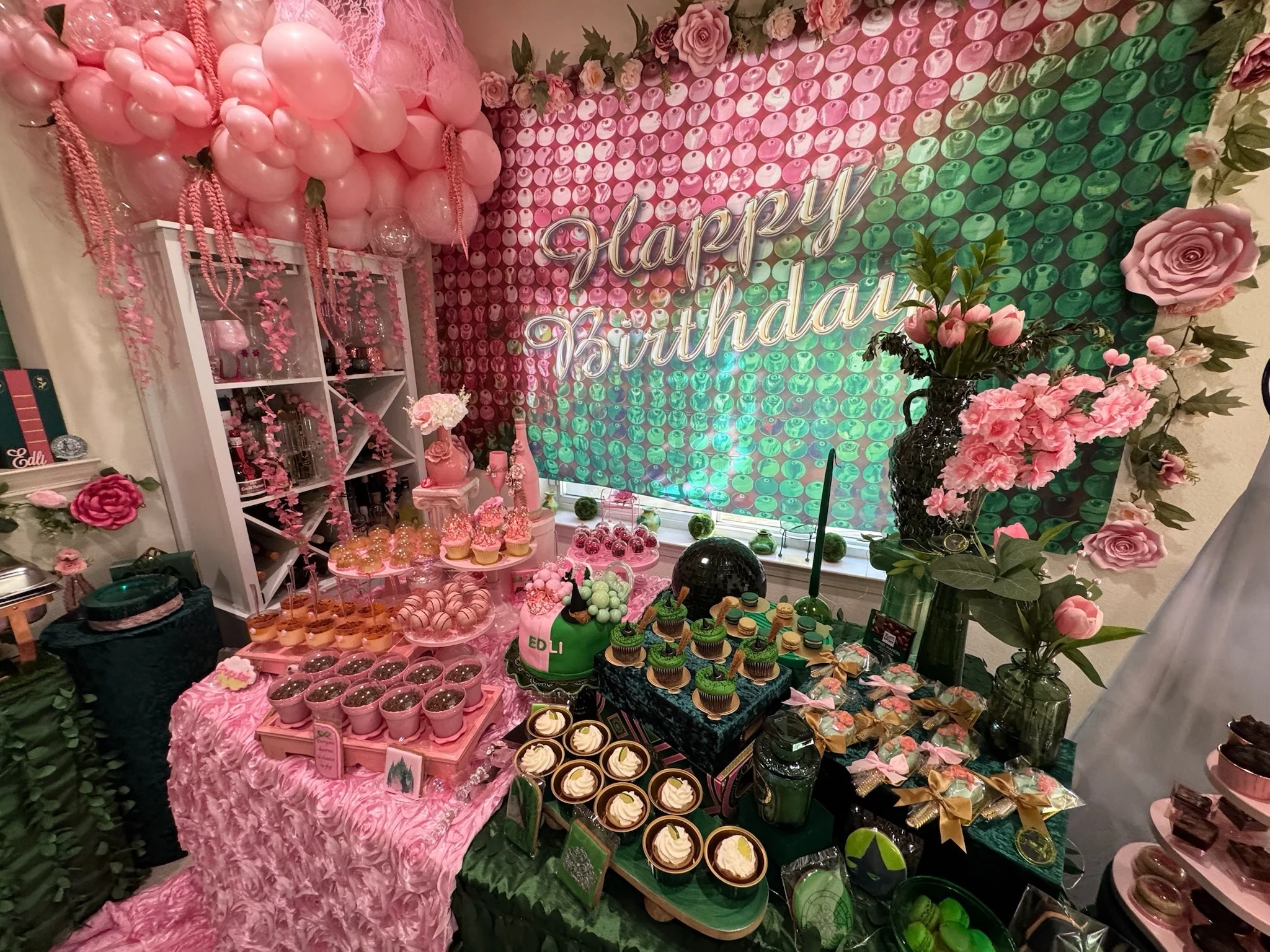 Lavish pink and green birthday dessert display with a Happy Birthday sign, balloon garland, tiered sweets, cupcakes, and oversized paper roses against a floral wall