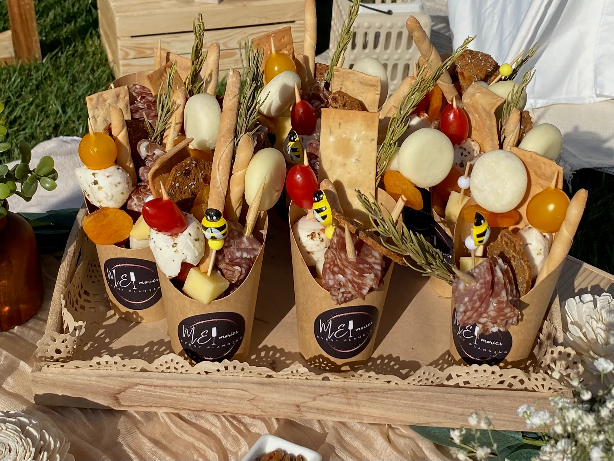 Individual charcuterie cones in kraft paper holders stamped with the Memories logo, filled with cured meats, aged cheeses, crackers, cherry tomatoes, and fresh rosemary