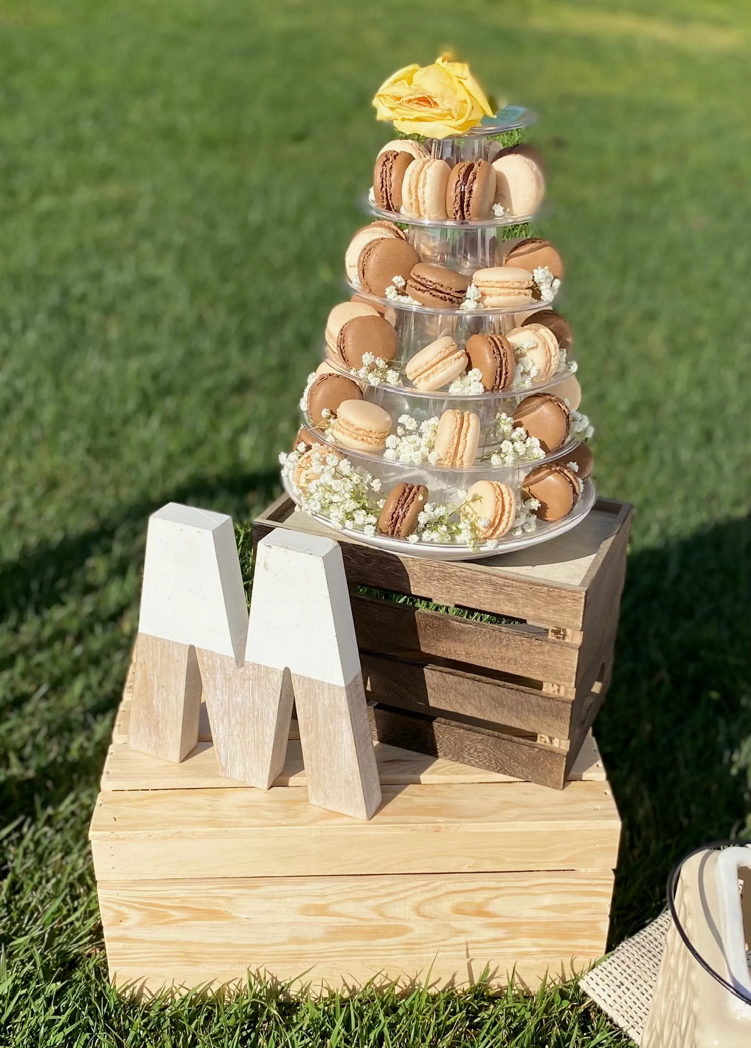 Tiered glass macaron tower with caramel and chocolate macarons decorated with baby breath flowers and topped with a yellow rose, displayed on stacked wooden crates