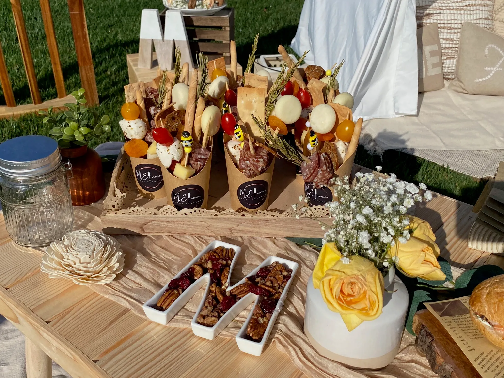 Rustic outdoor catering table with branded charcuterie cones, small bowls of pecans and dried fruit, a vase of yellow roses and baby breath, and a wooden M letter decoration