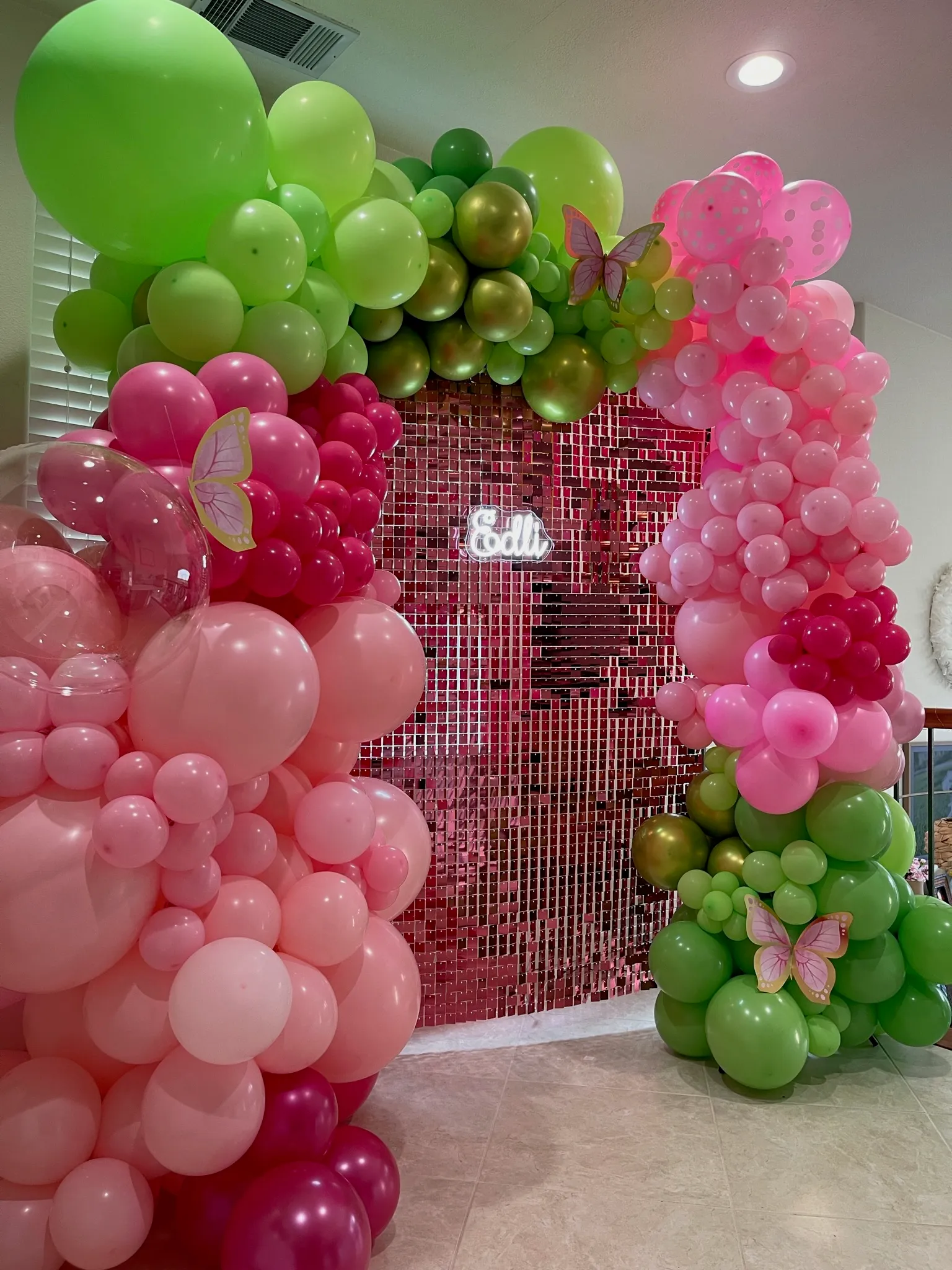 Vibrant pink and green balloon arch framing a rose gold sequin shimmer wall, decorated with butterfly accents and a neon name sign for a birthday party