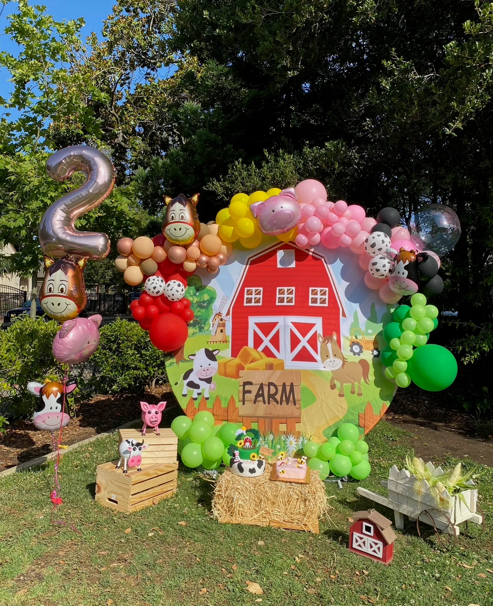 Outdoor farm-themed second birthday party with a red barn backdrop, colorful balloon garland in pink, yellow, green, and brown, animal cutouts, hay bales, and a themed dessert table