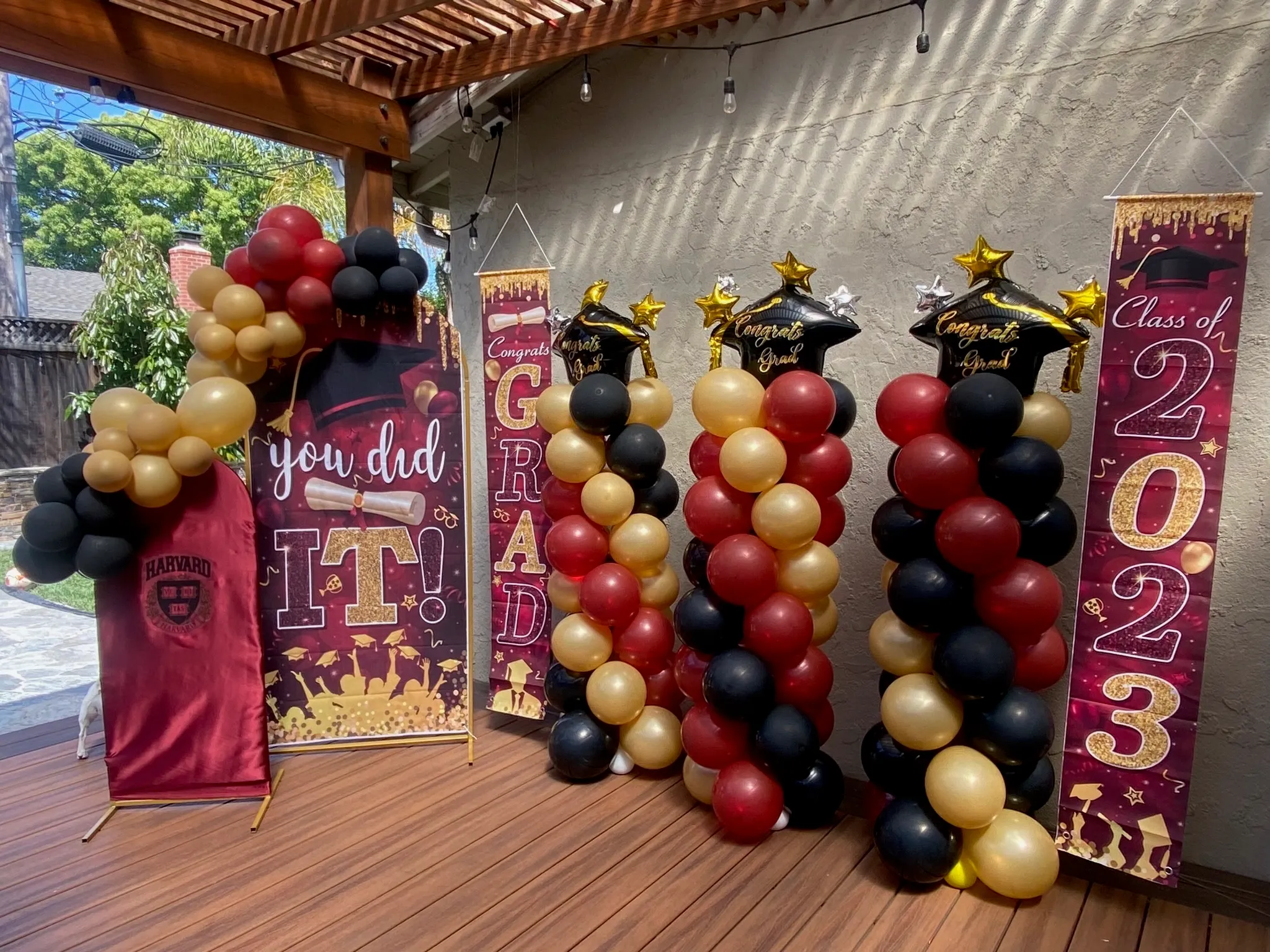 Harvard-themed graduation celebration with maroon, black, and gold balloon columns spelling GRAD, star-shaped balloons, and Class of 2023 banners on a covered patio