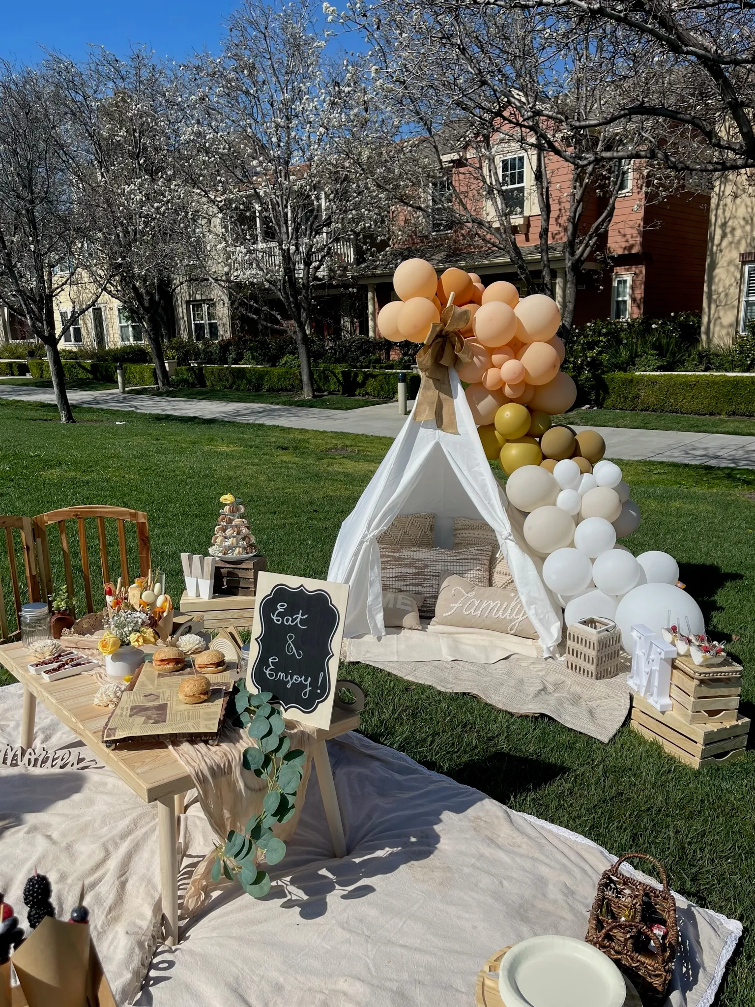 Close-up of an elegant outdoor picnic with a white teepee tent, warm-toned balloon garland, chalkboard Eat and Enjoy sign, and a rustic food table surrounded by eucalyptus greenery