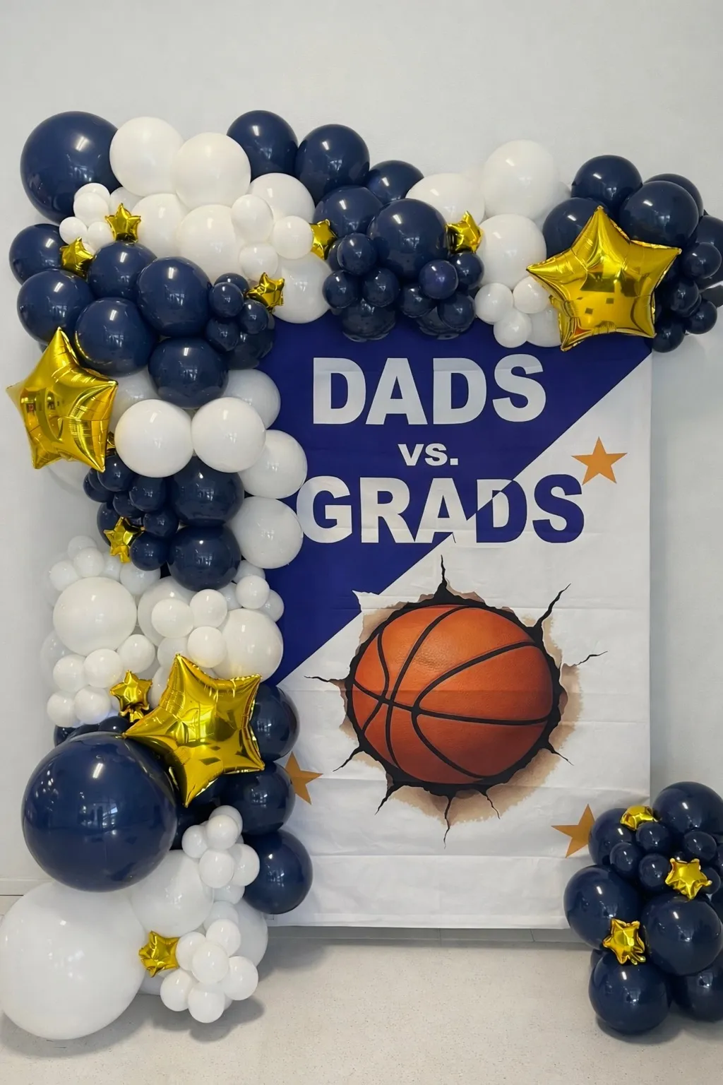 Custom navy blue and white balloon garland installation framing a Dads vs Grads basketball-themed event poster with gold star accent balloons