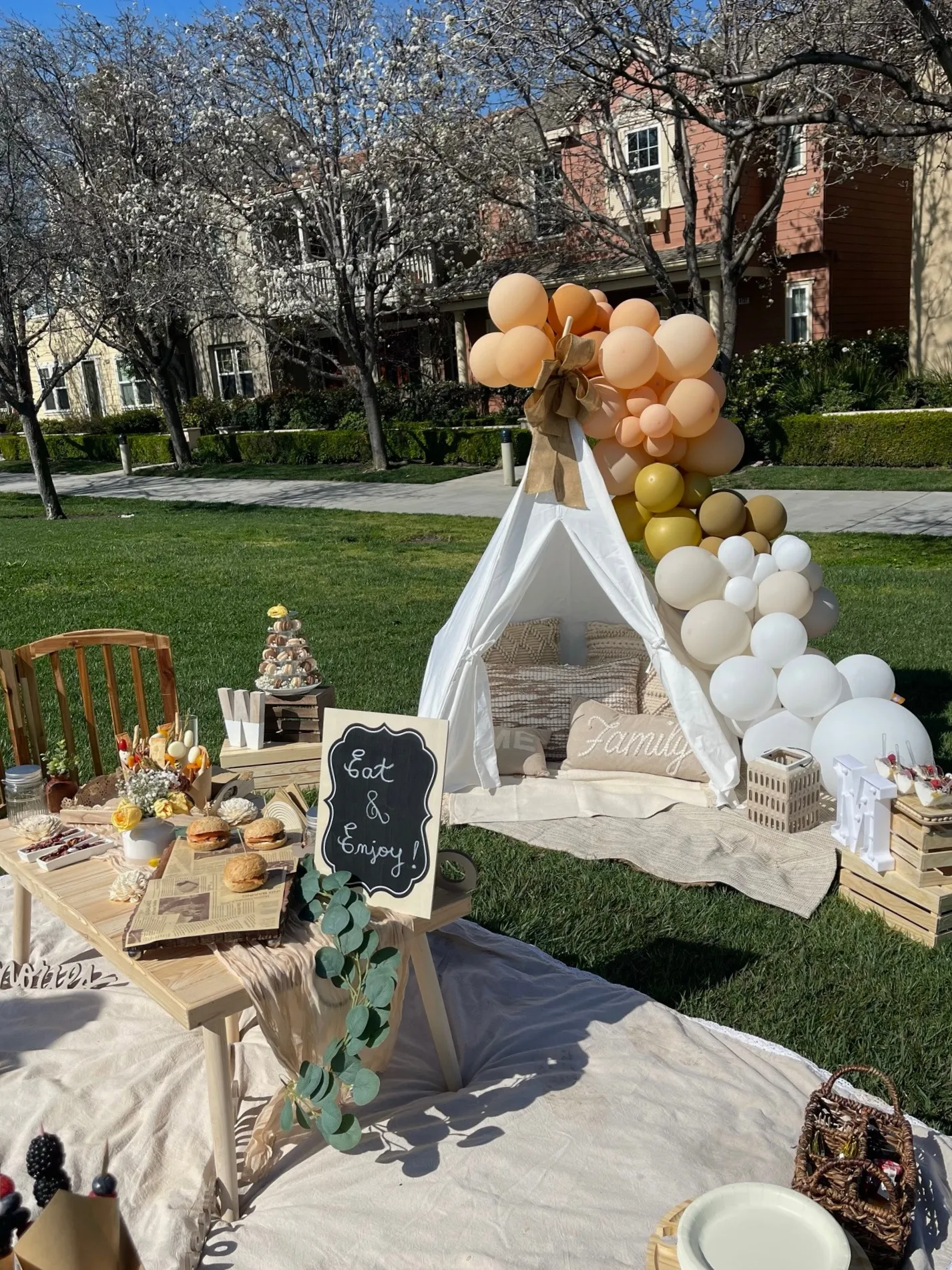 Elegant outdoor picnic celebration with a white teepee tent, warm-toned balloon garland in peach and gold, a styled food table, eucalyptus garland, and an Eat and Enjoy chalkboard sign set under blooming cherry trees