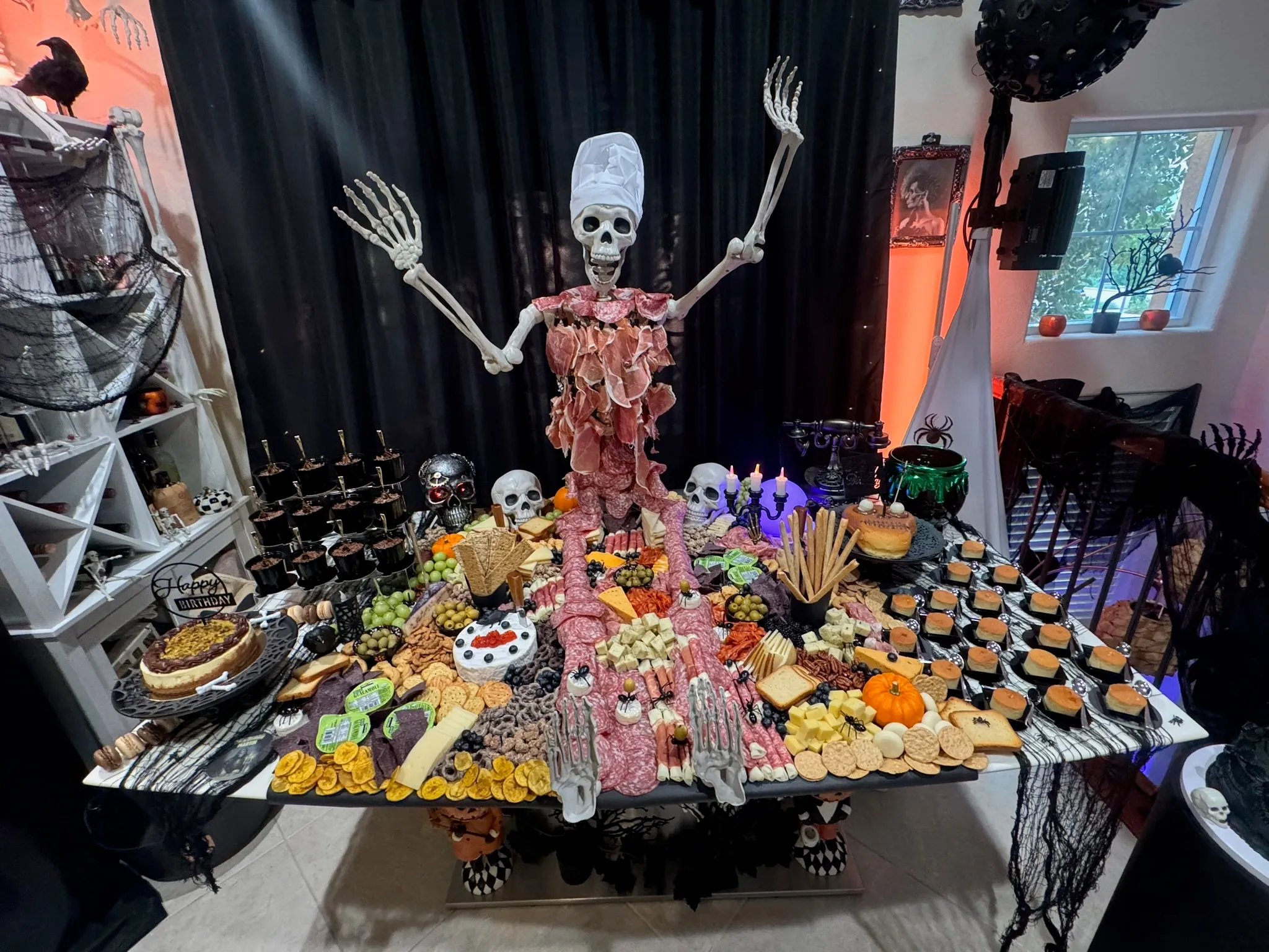 Elaborate Halloween-themed styled table featuring a skeleton centerpiece draped in charcuterie, surrounded by themed desserts, cupcakes, cauldron props, and spiderweb decorations